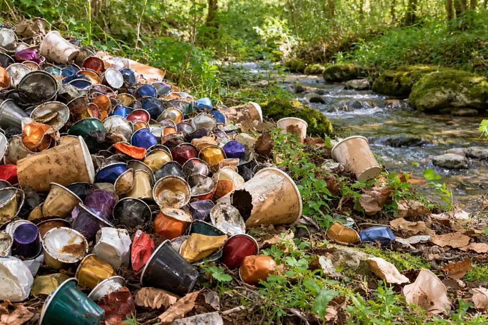 Un monticule de capsules de café usagées de toutes les couleurs et de gobelets jetables, déversé dans une clairière au bord d'un ruisseau.